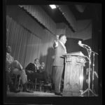 Dr. Martin Luther King, Jr. speaks at a podium. Three men sit behind him.