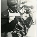 George Washington Carver holds crocheting needles and crochets yarn into a garment