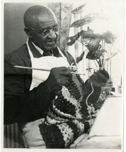 George Washington Carver holds crocheting needles and crochets yarn into a garment